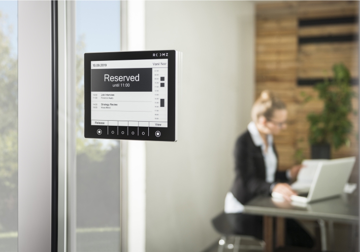 Woman in a meeting room - ROOMZ display panel shows a meeting in in progress